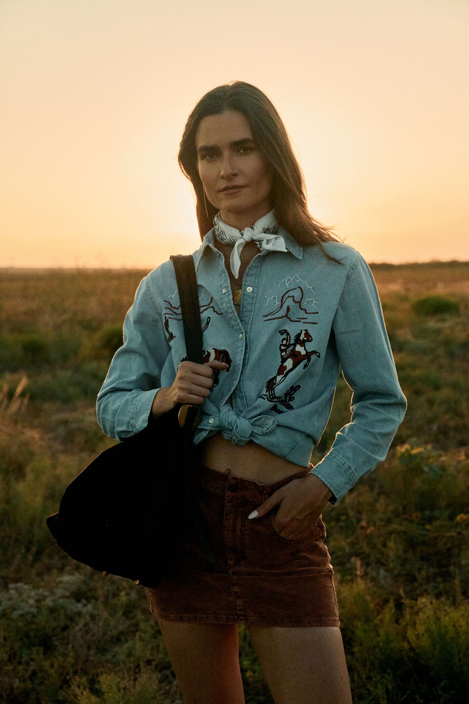 Woman in embroidered denim jacket and brown shorts holding a bag at golden hour in an open field.
