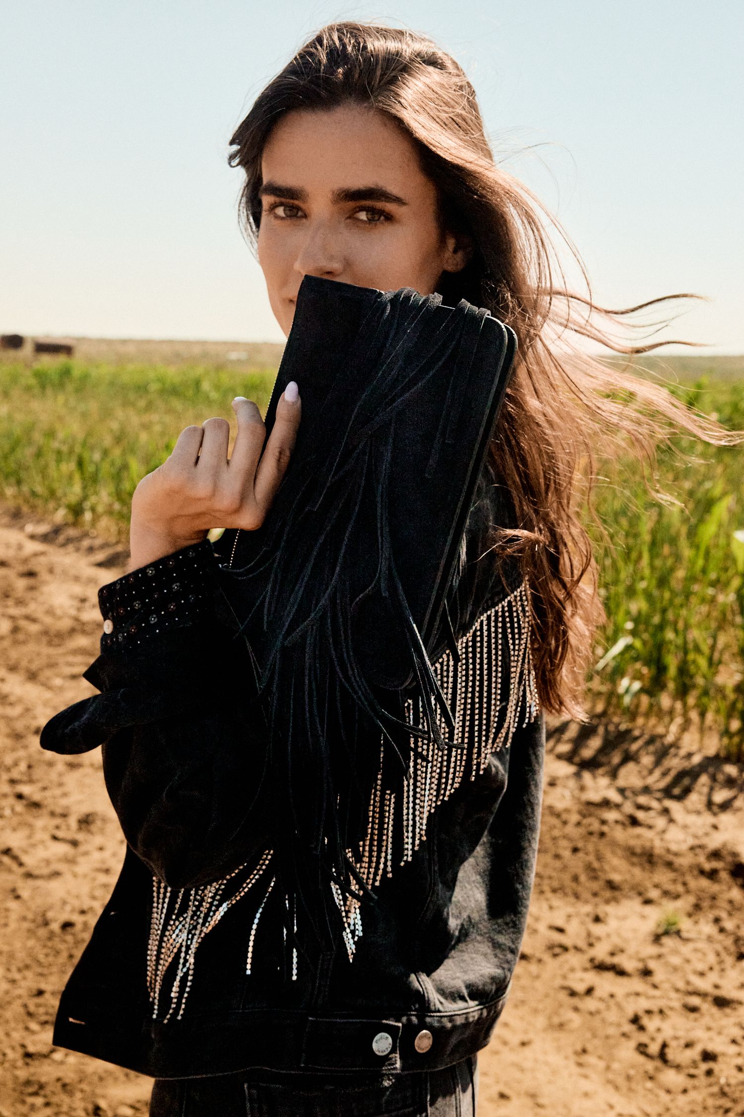 Brunette model in black studded fringe jacket holds sleeve to face in a sunny agricultural field.