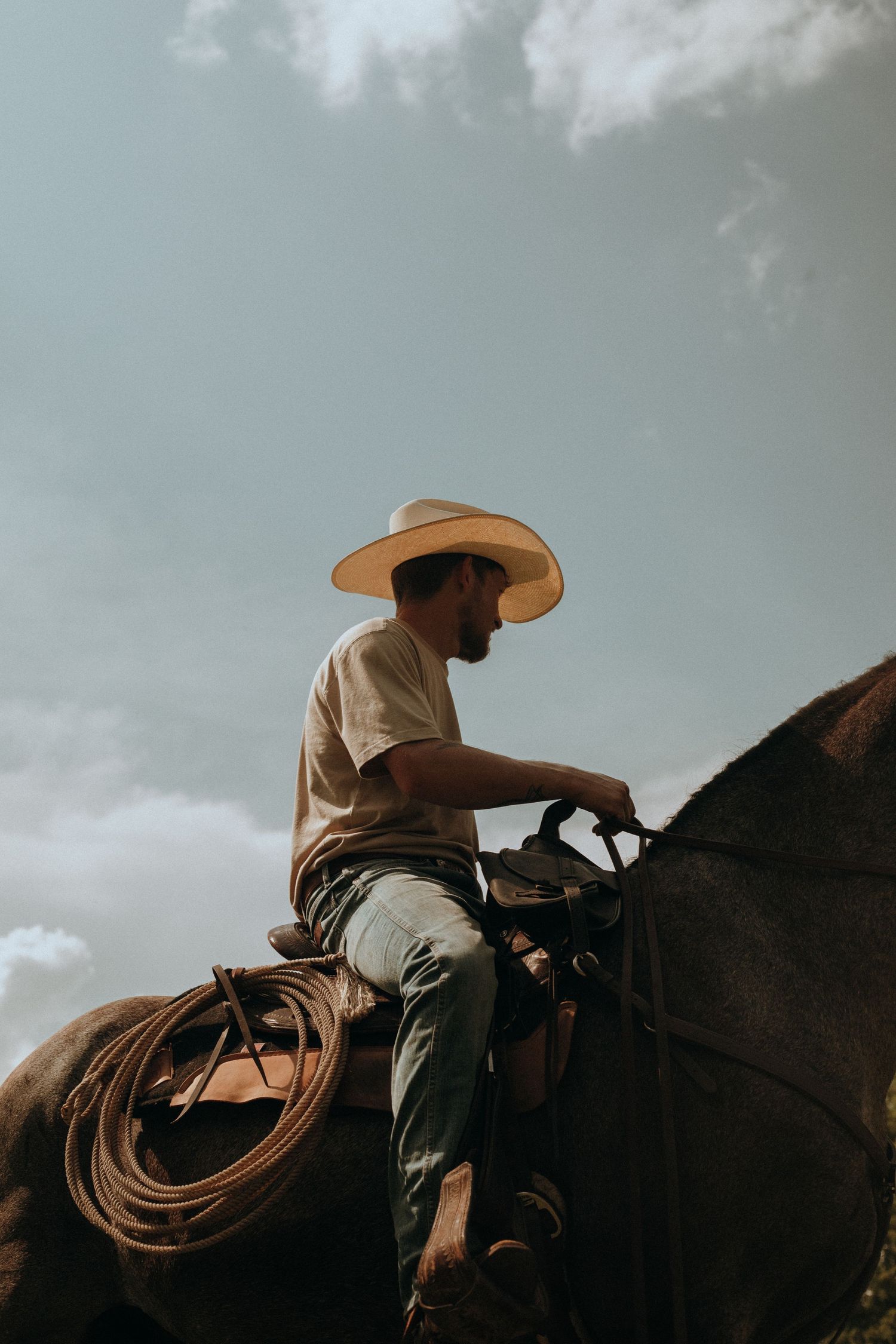 Low angle view of a cowboy in white hat sitting on horseback against a cloudy sky with rope on saddle.