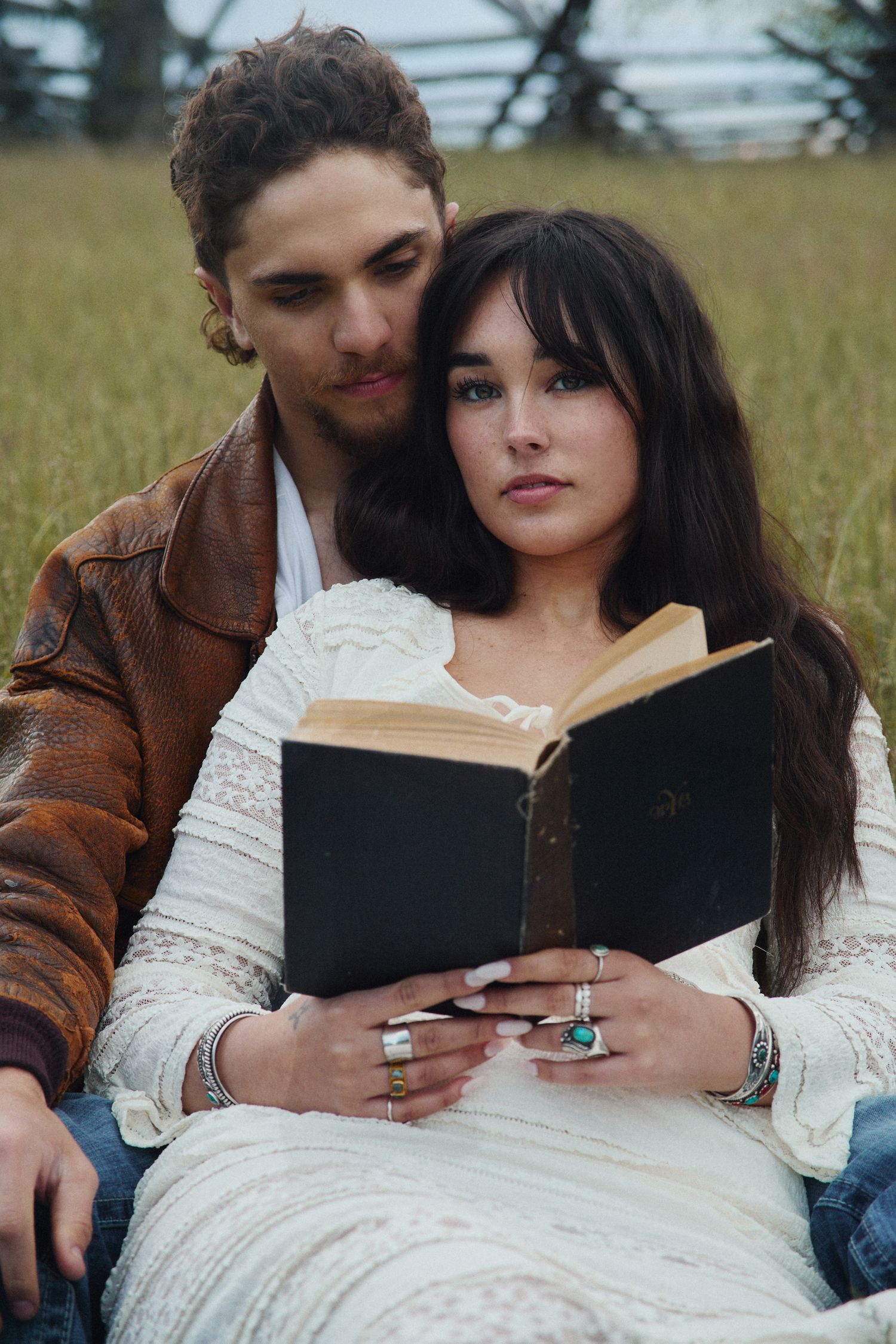 Couple sitting in a field, woman in white lace dress holding a book while man in brown leather jacket embraces her.