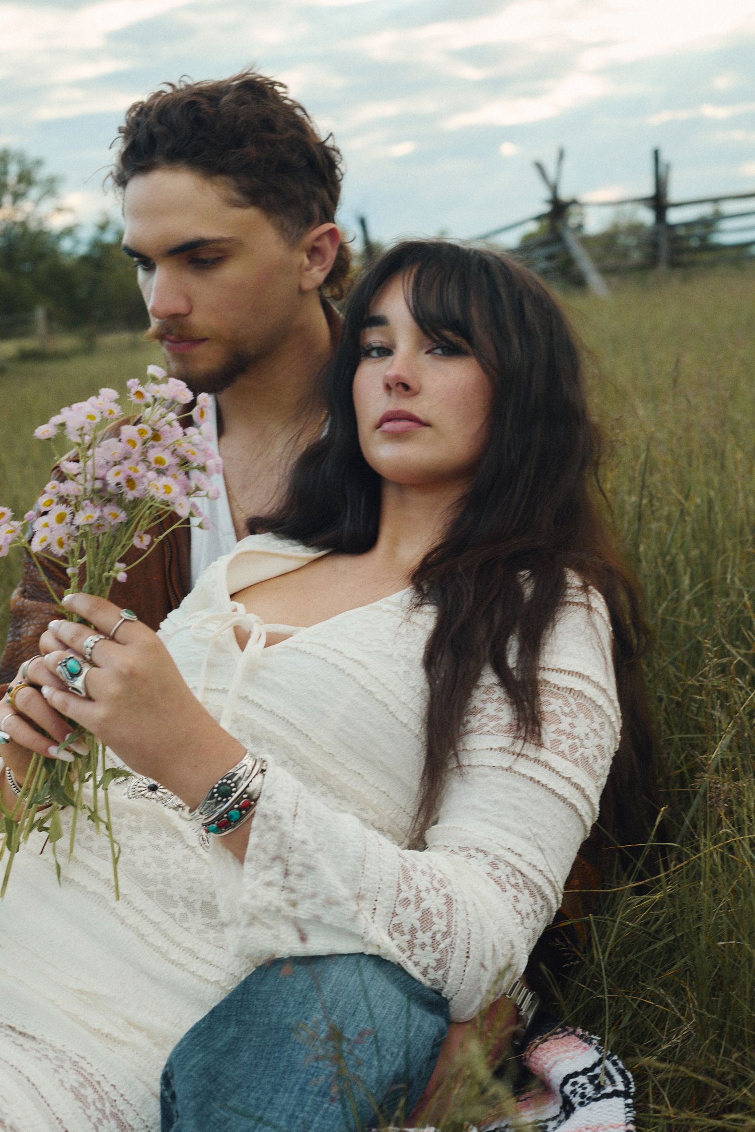 A bohemian couple sits in a field, woman in white lace dress holding wildflowers, leaning against a man outdoors.