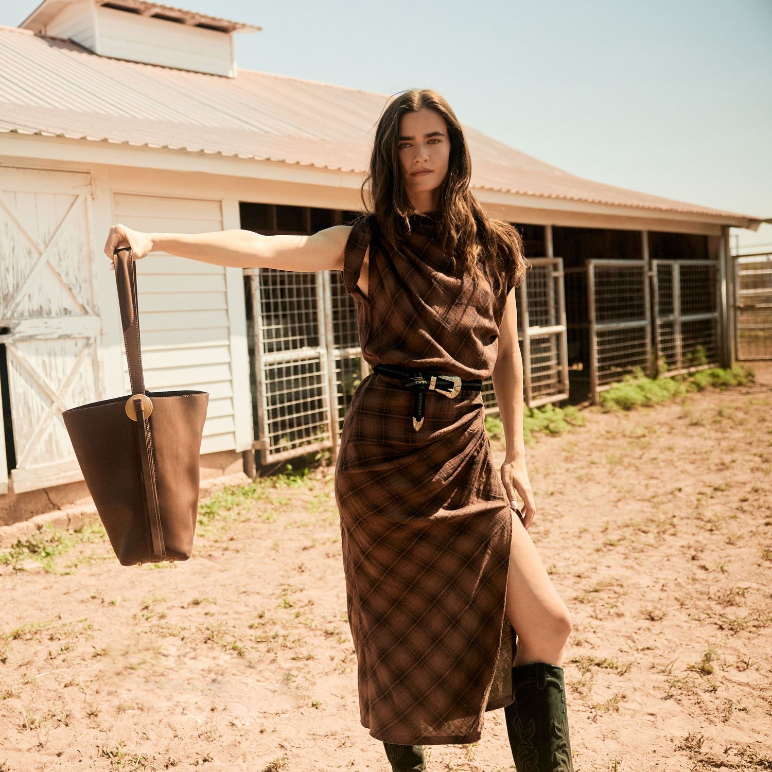 Woman in plaid midi dress with belt and green boots holds a brown leather tote bag at a sunny ranch.