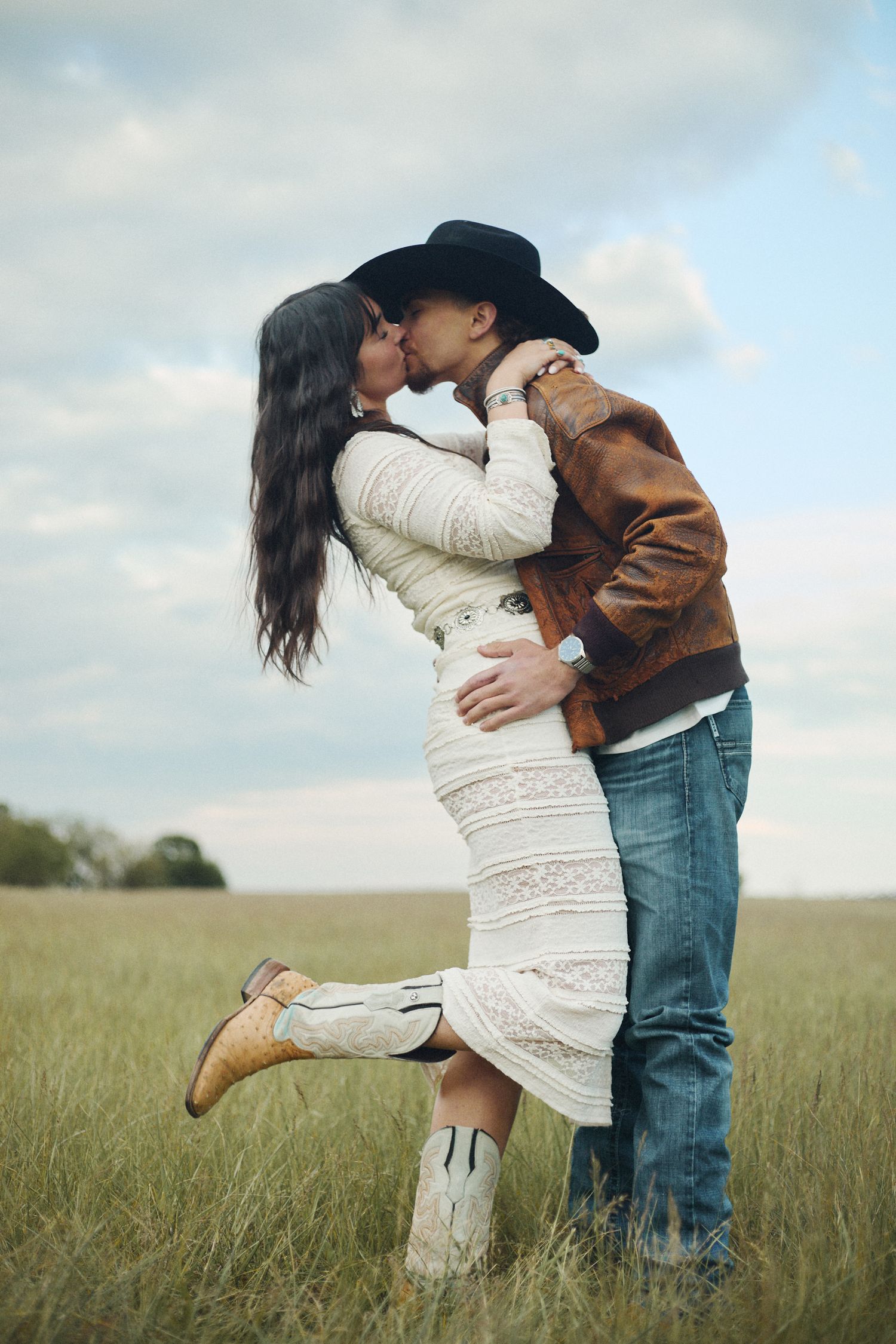 Couple kissing in a grassy field, woman in white lace dress and cowboy boots, man in brown leather jacket and black hat.