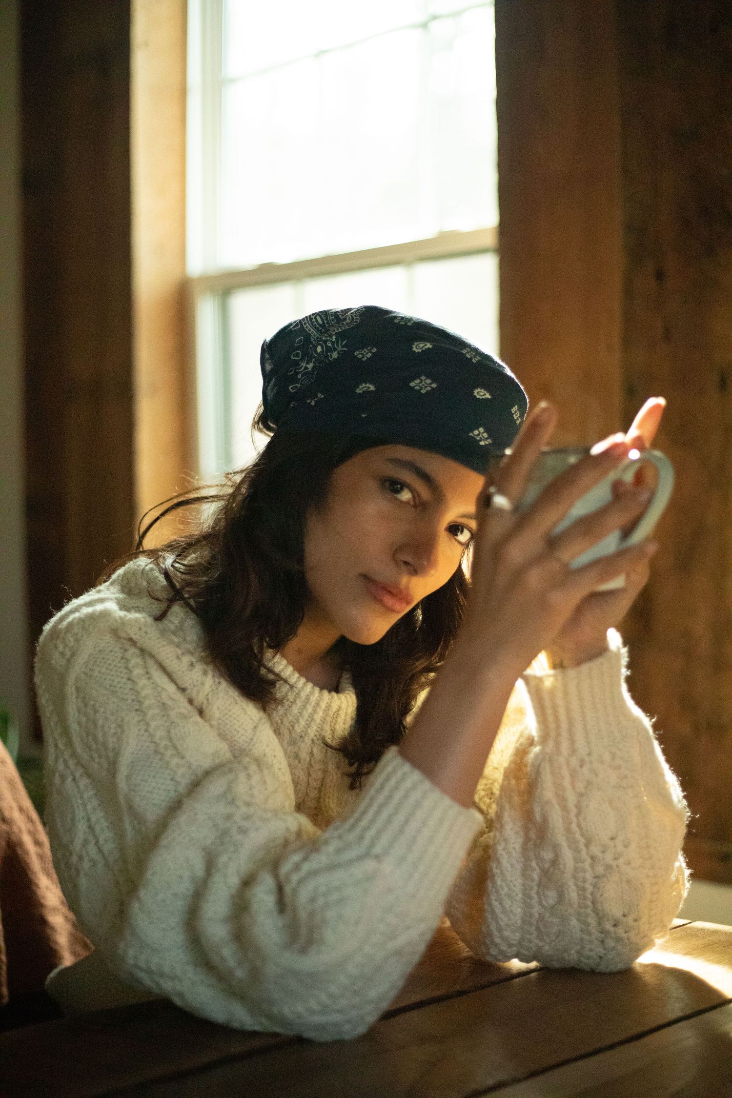 Young woman in white knit sweater and bandana headscarf holds mug by window.
