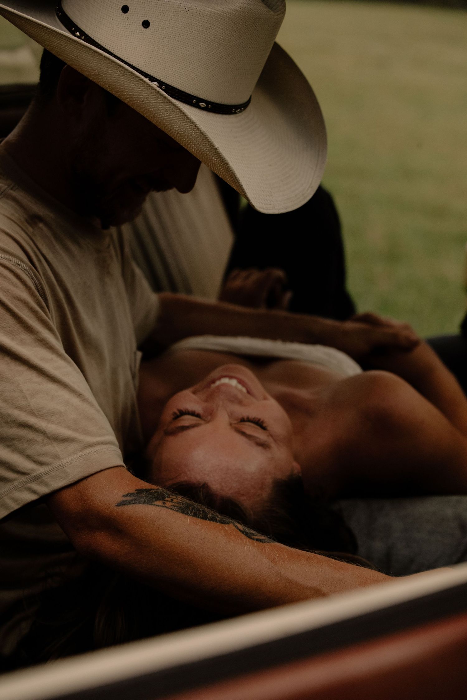 Intimate moment of a cowboy in white hat leaning over a woman lying back smiling outdoors.