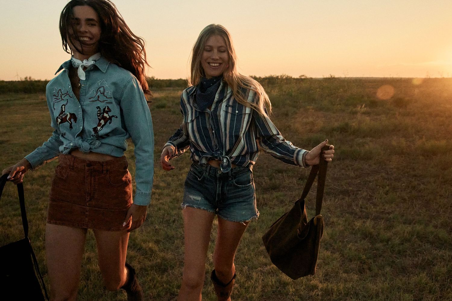 Two smiling women walking through a golden field at sunset, wearing western-style shirts and shorts.