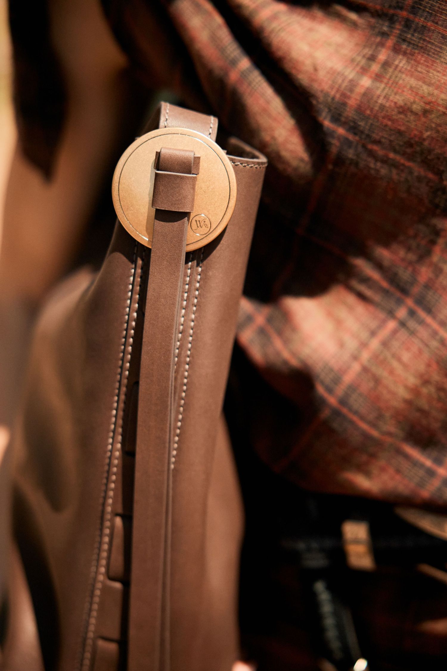 Close-up of a brown leather bag with circular buckle detail against plaid fabric.