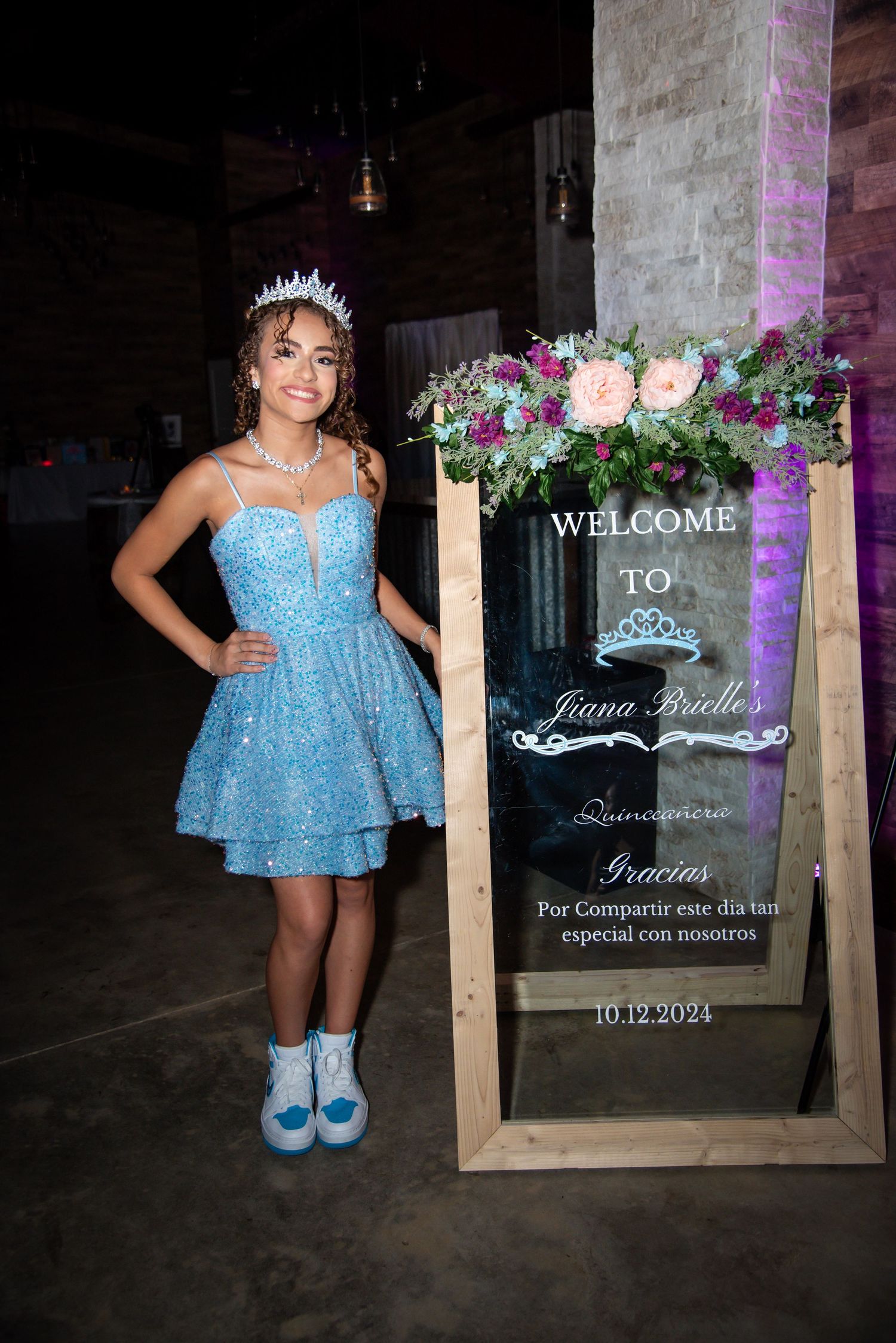 Young woman in blue sequin dress and tiara poses beside a floral quinceañera welcome sign dated 10.12.2024.