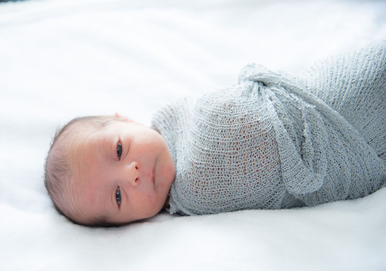 Newborn baby swaddled in a soft gray knit wrap lying peacefully on a white blanket.