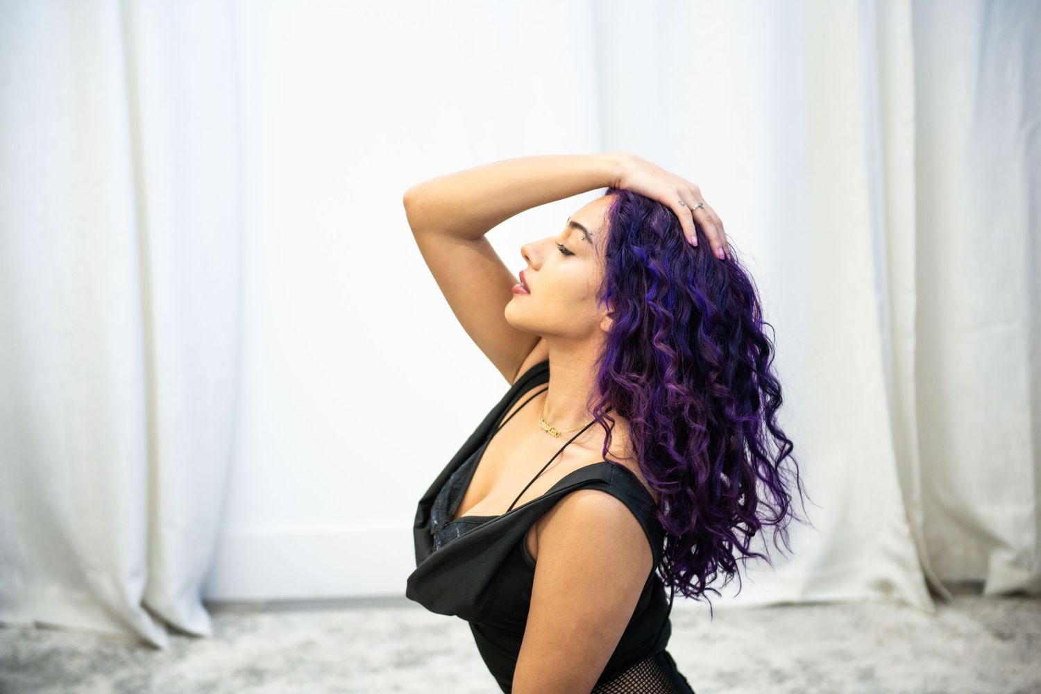 Woman with vibrant purple curly hair poses gracefully in black top against white curtain backdrop.