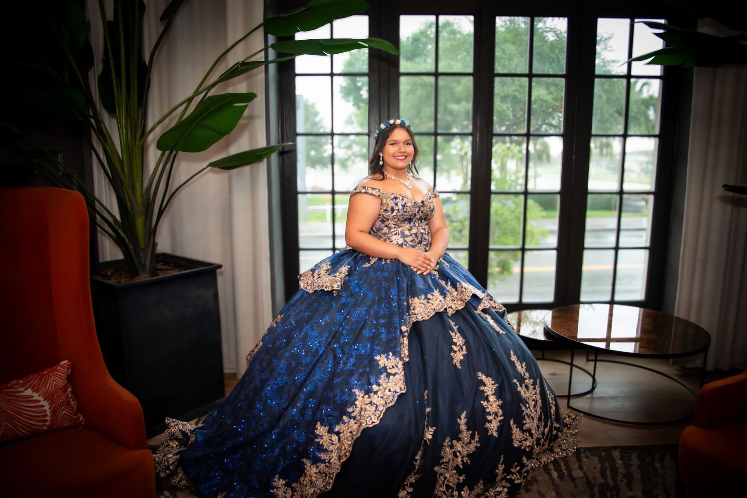 Young woman in elegant navy blue and gold quinceañera ball gown posing indoors near large windows.