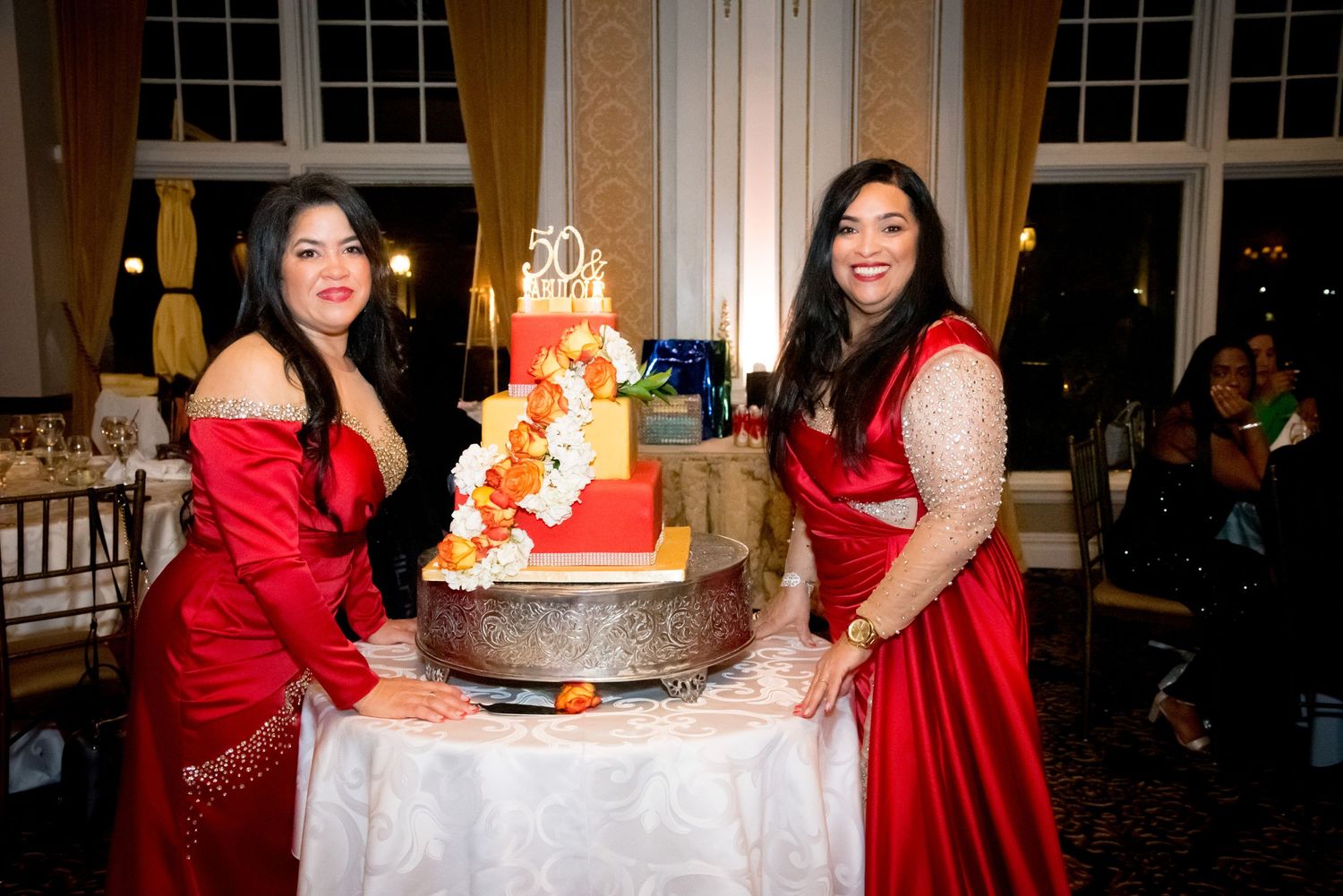 Two women in red dresses pose with elegant tiered wedding cake.