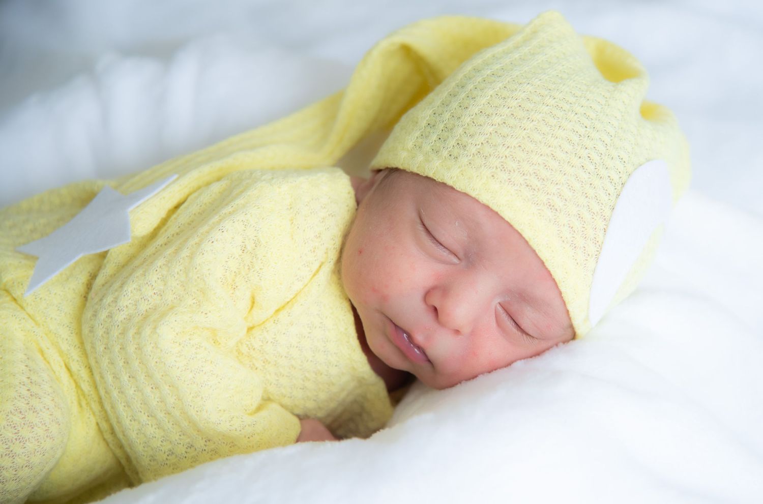Newborn baby sleeping peacefully wrapped in a soft yellow knit blanket and matching hat on white bedding.