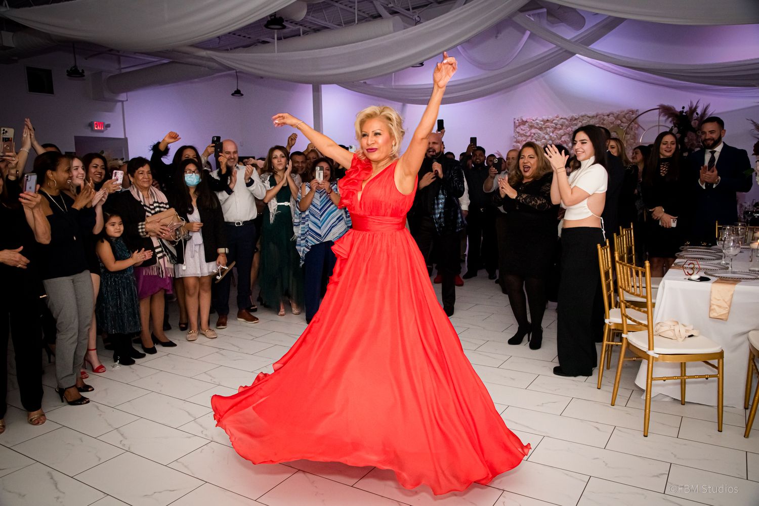 Woman in red gown dancing at elegant event with guests applauding in background.