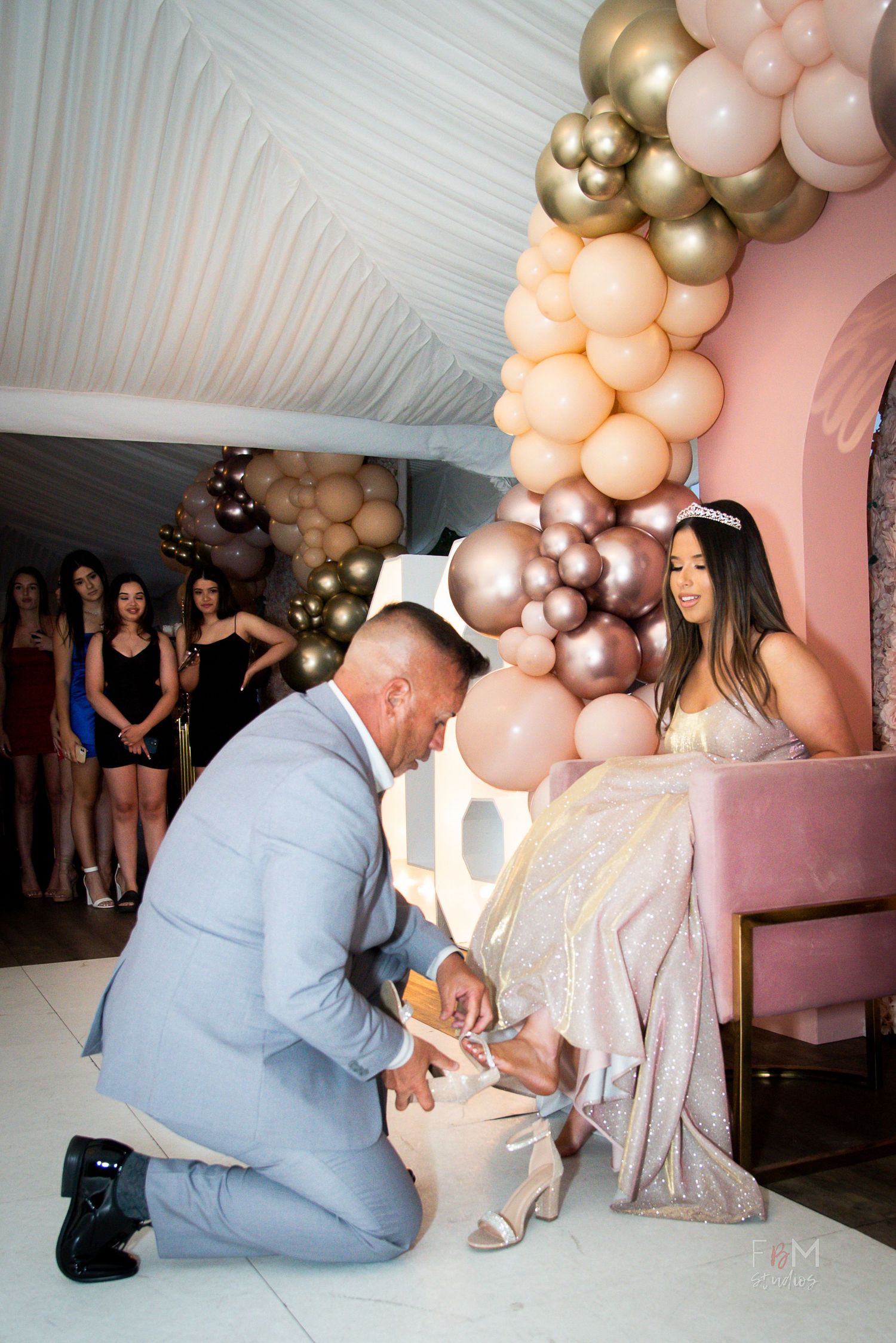 Man in gray suit kneeling to put heels on a woman in sequin dress at elegant quinceañera with balloon arch by FBM Studios in Fort Lauderdale