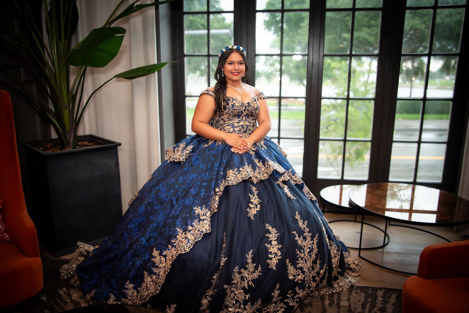 Young woman in navy blue and gold quinceañera ball gown poses elegantly indoors near large windows by FBM Studios in Fort Lauderdale Florida