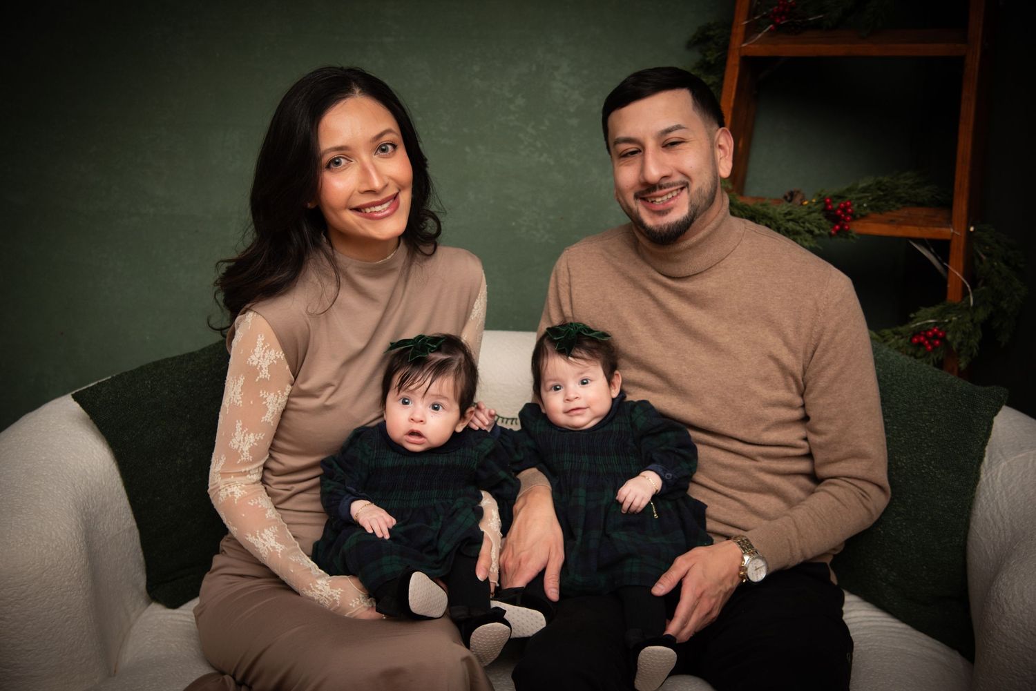 A smiling family of four with twin babies in black outfits poses on a couch against a green backdrop.