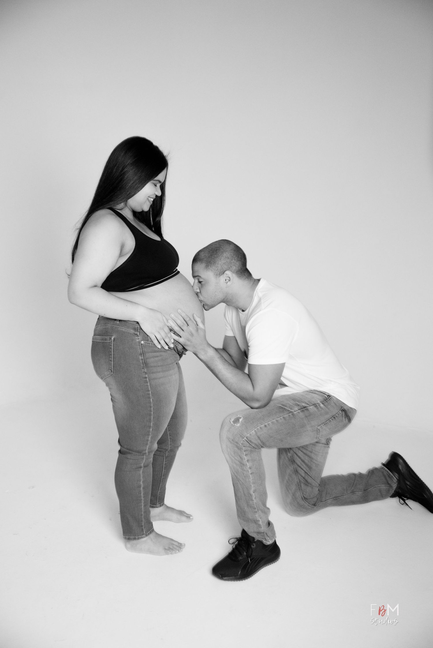 Black and white maternity photo of a man kneeling and kissing pregnant womans belly in a studio setting.