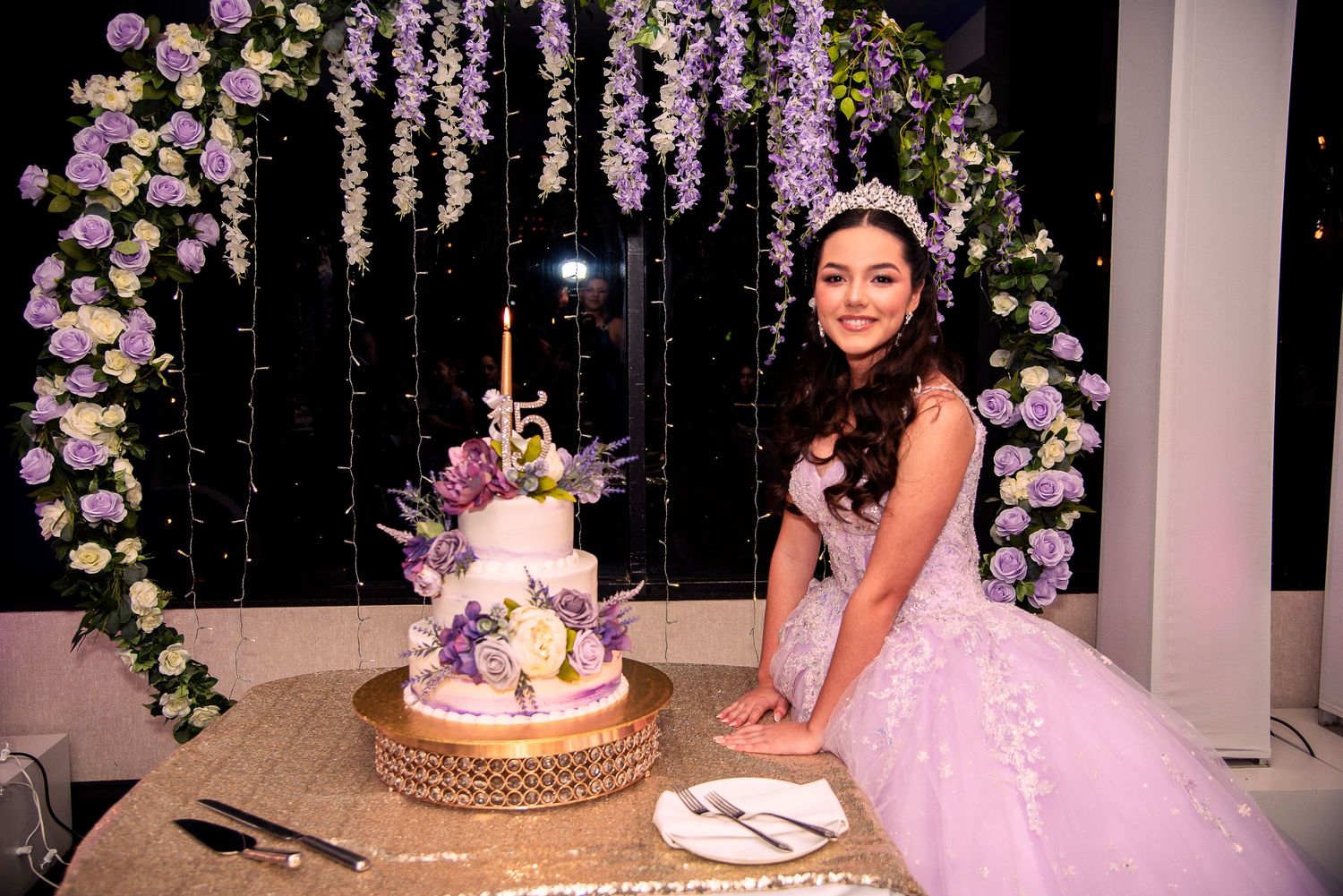 Young woman in pink quinceañera gown poses beside a purple floral decorated cake at her 15th birthday celebration by FBM Studios in Fort Lau