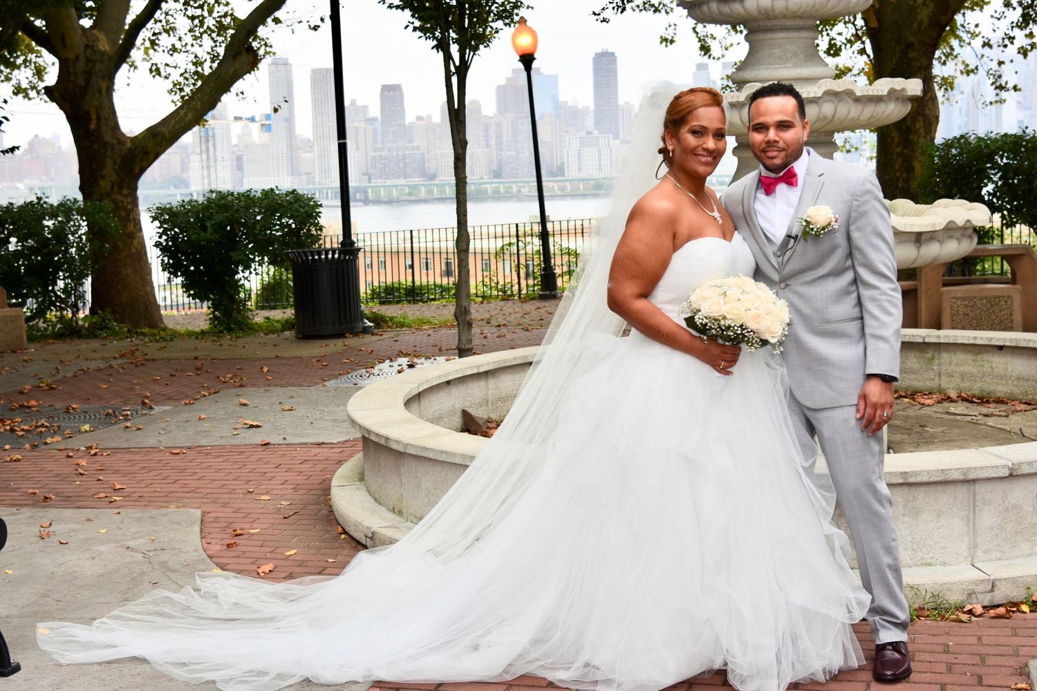 A bride and groom pose for wedding photos with the NYC skyline visible in the background at Liberty State Park by FBM Studios in Fort Lauder