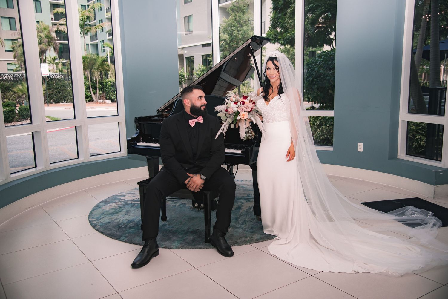 Bride in white gown and groom in black tuxedo pose by a grand piano in an elegant sunlit room.