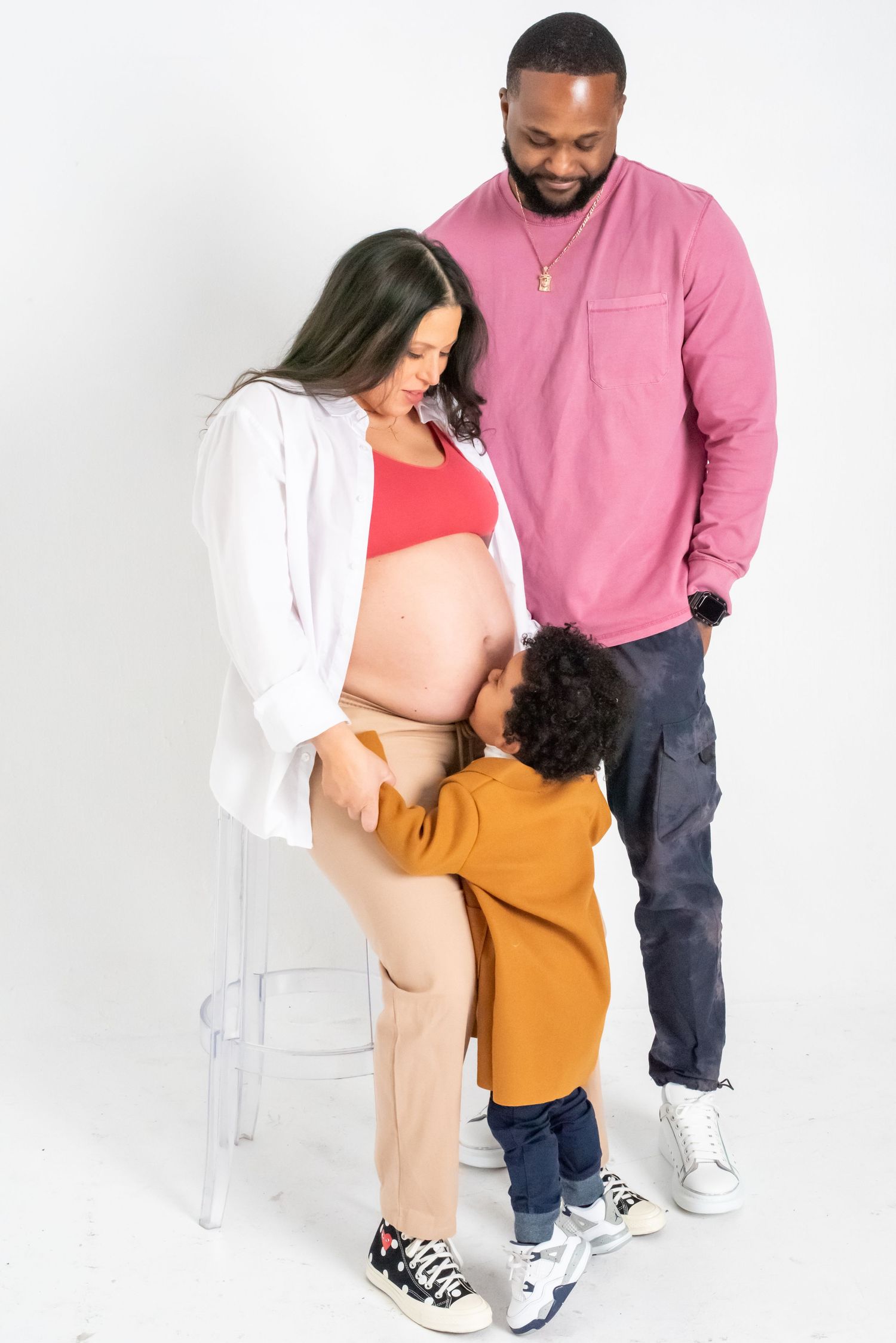 Pregnant woman with toddler kissing her belly, standing beside a man in a pink shirt during a maternity photoshoot.