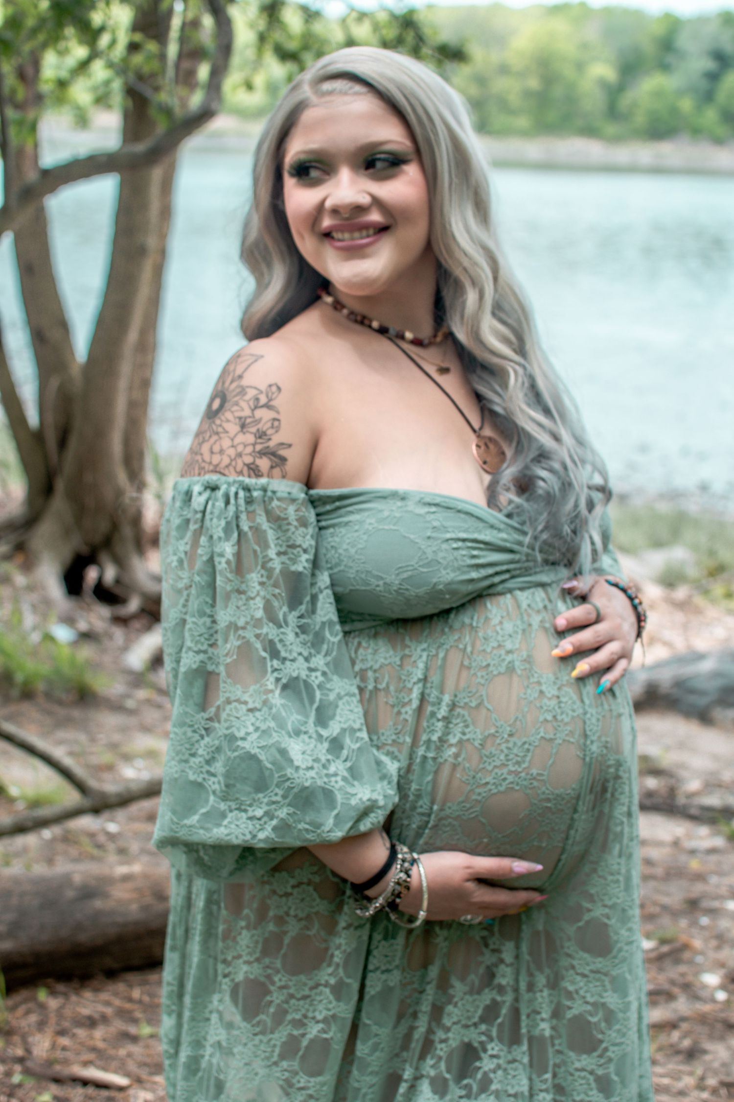 Pregnant woman in sage green lace off-shoulder maternity gown smiling outdoors near a lake with gray hair and tattoos.