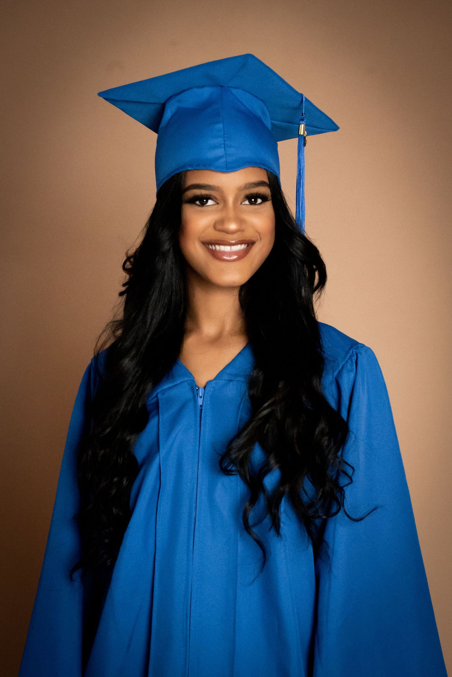 Smiling young woman in blue graduation cap and gown posing for a portrait photo against a tan background.