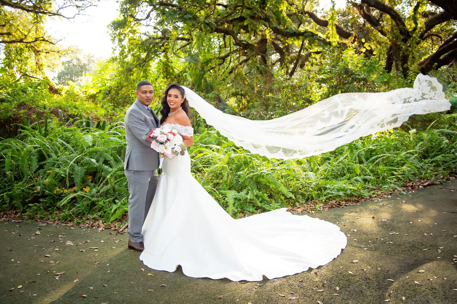 Bride and groom pose in lush garden with flowing veil billowing in the wind on their wedding day.
