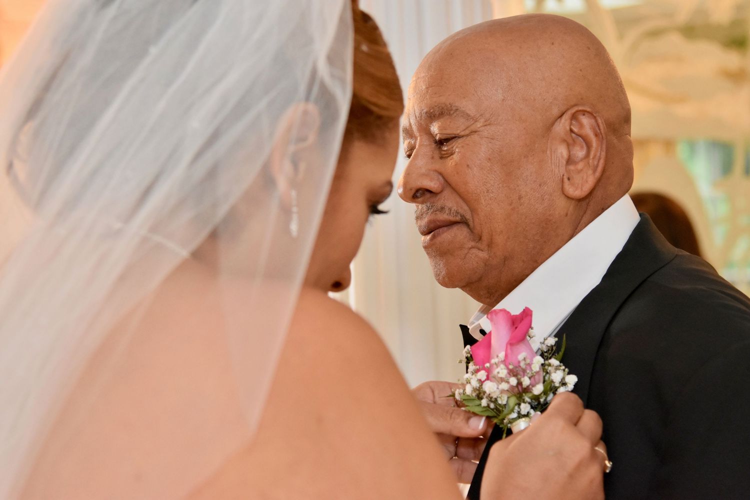 Emotional father-daughter wedding moment, elderly man in tuxedo with pink boutonniere embracing bride in white veil.