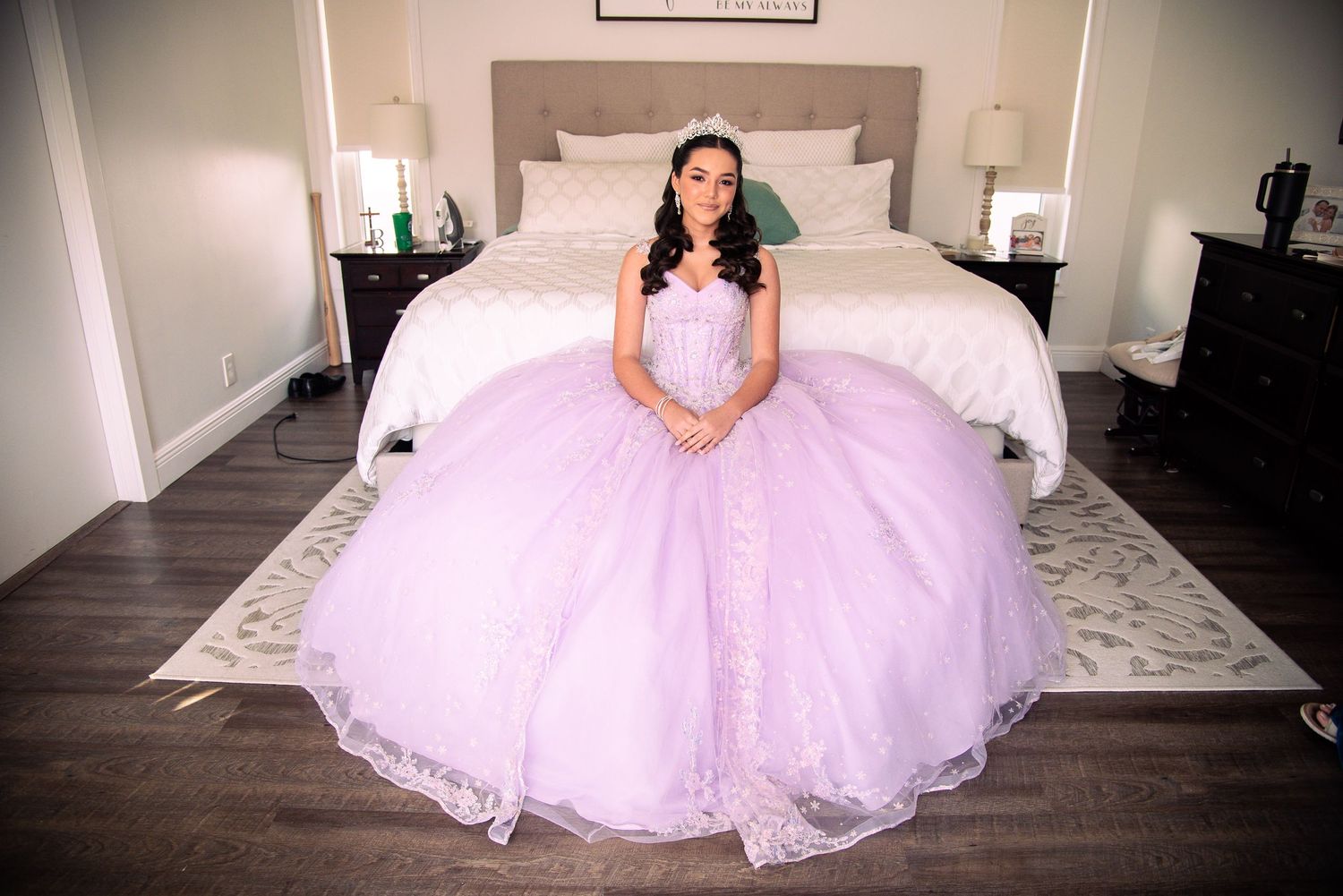 Young woman in lavish lavender quinceañera ball gown sitting gracefully on bed, wearing tiara in elegant bedroom setting.