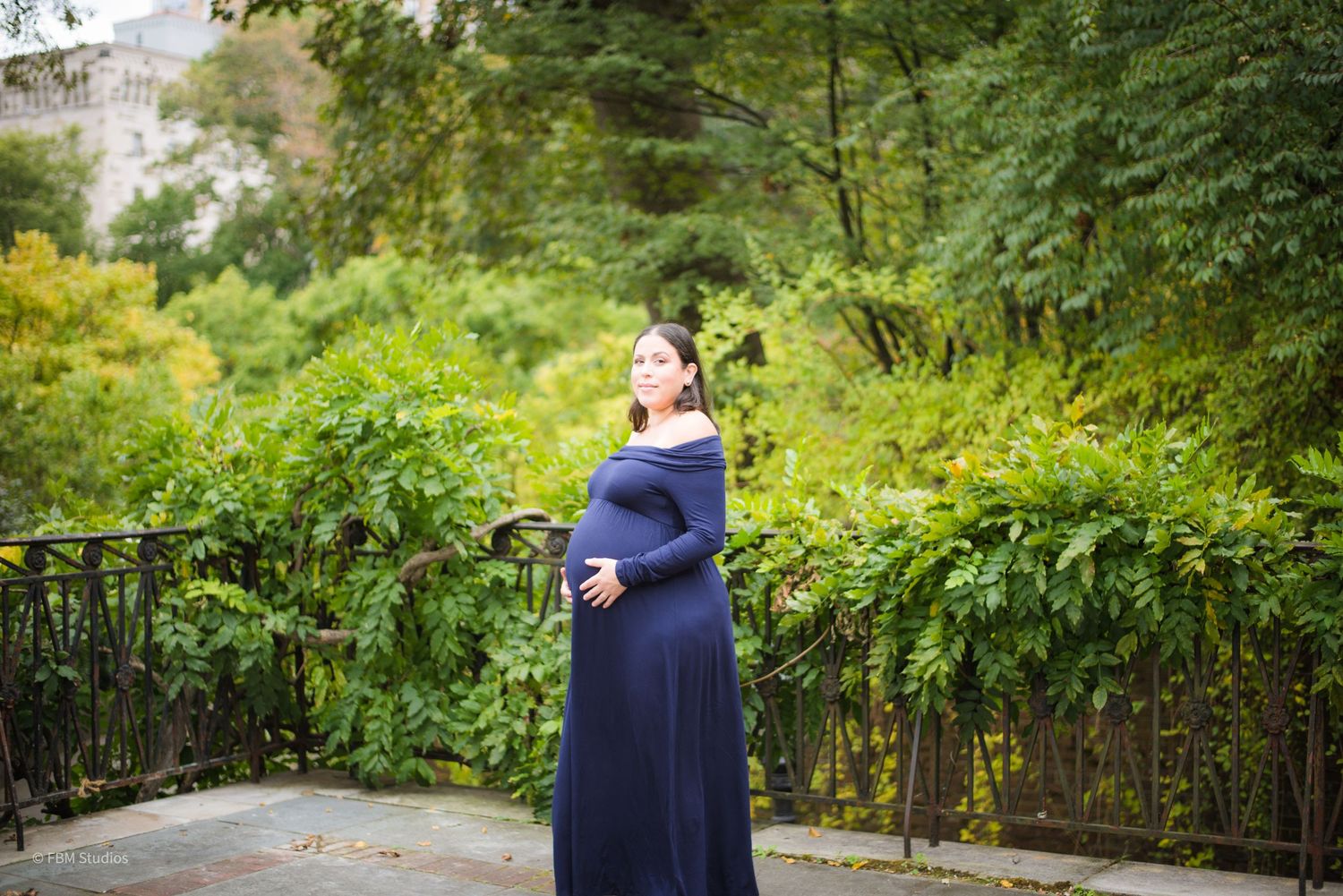 Pregnant woman in navy blue maxi dress posing outdoors in lush green park setting during maternity photo session.