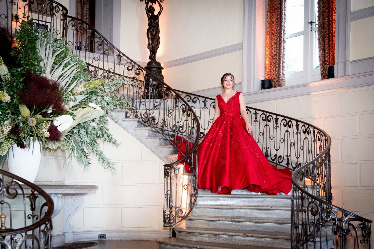 Woman in elegant red ball gown poses on ornate spiral staircase adorned with lush floral arrangements in grand venue by FBM Studios in FL