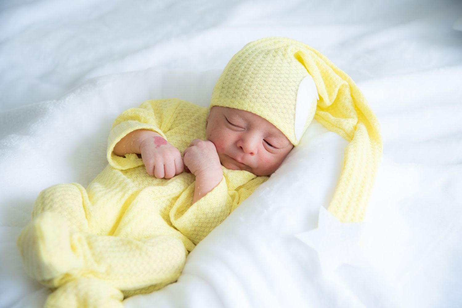 Newborn baby sleeping peacefully in yellow knit outfit and hat on white fluffy blanket.