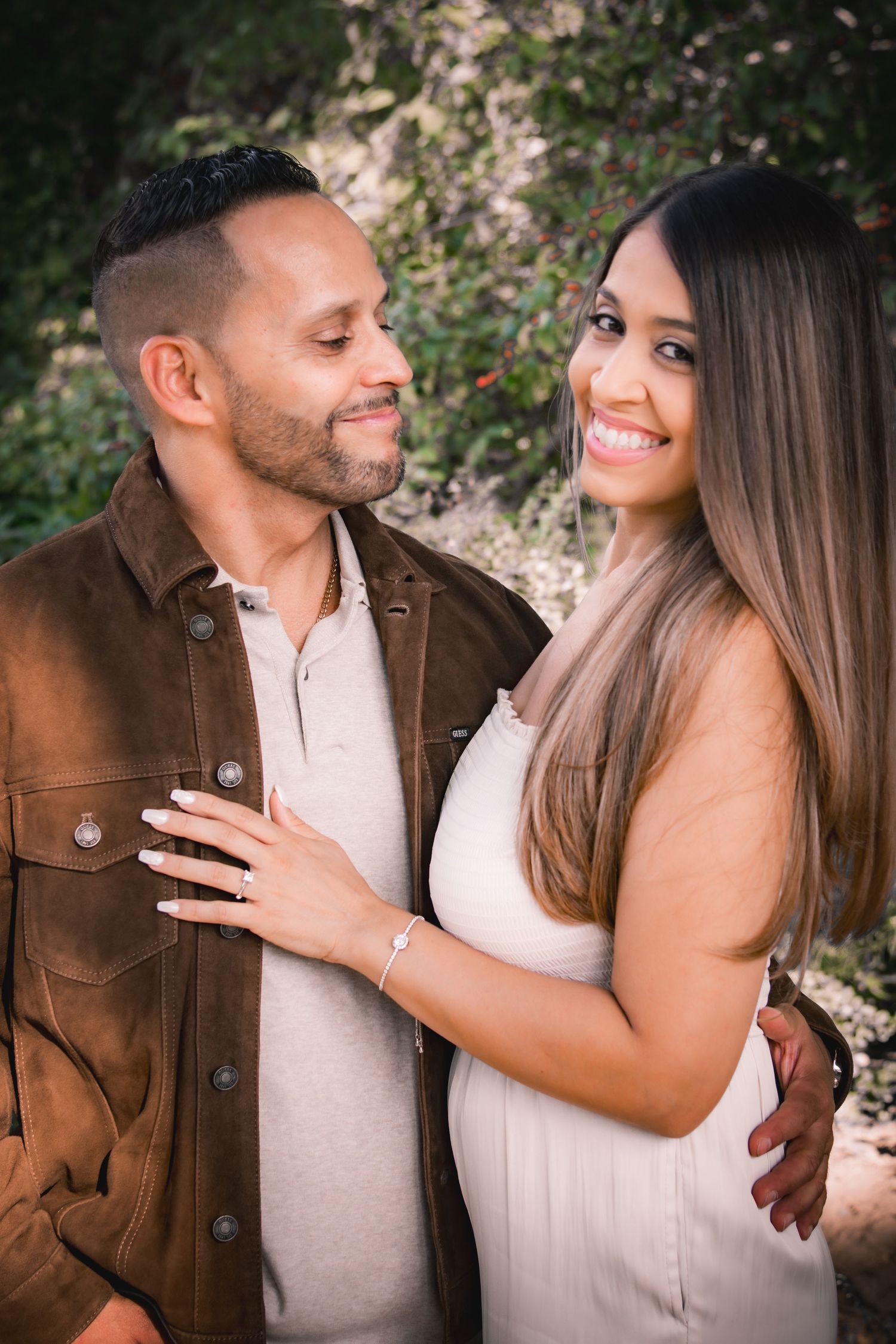 A smiling couple poses outdoors, woman in white dress with engagement ring, man in brown jacket, surrounded by greenery.