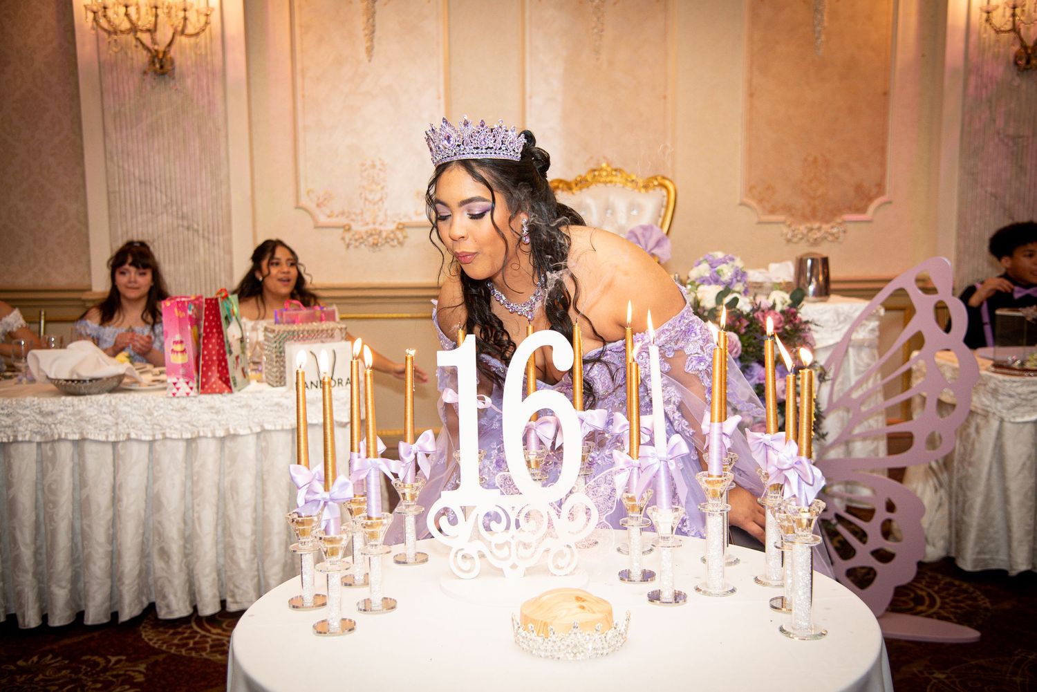 A young woman in a tiara blows out candles on a Sweet 16 celebration table at an elegant party venue.