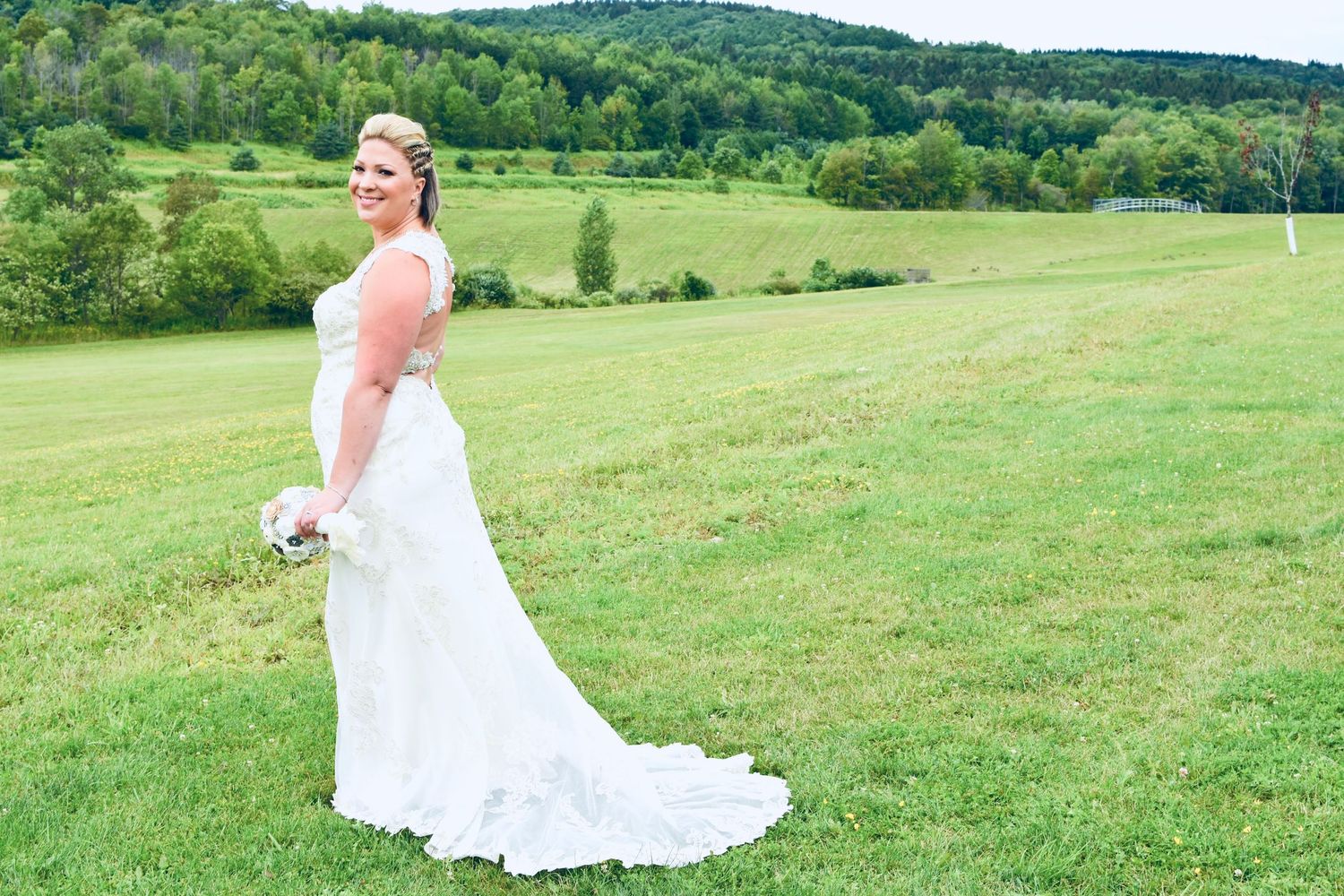 Bride in white lace gown with train poses on green meadow, smiling over shoulder with bouquet, rolling hills behind.
