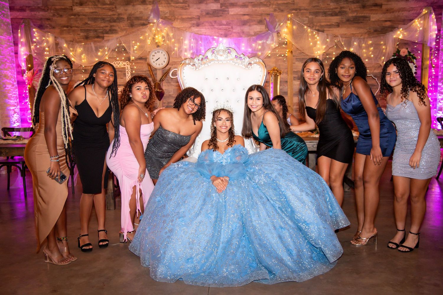 A quinceañera girl in a blue ball gown poses with her court of friends at a decorated celebration venue.