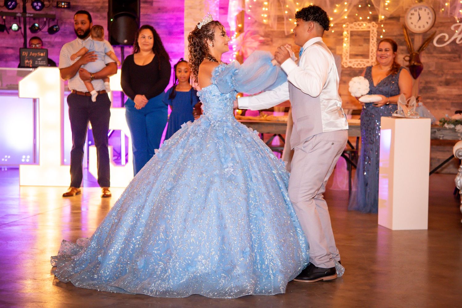 A young woman in a sparkling blue ball gown dances with a partner at a quinceañera celebration.