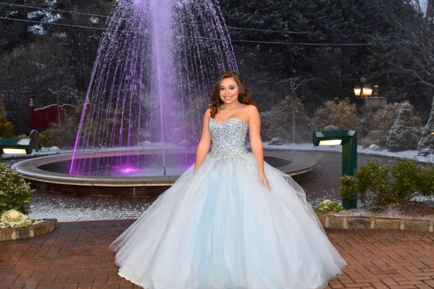A person in a white ballgown poses in front of an illuminated purple fountain during a nighttime photo session.