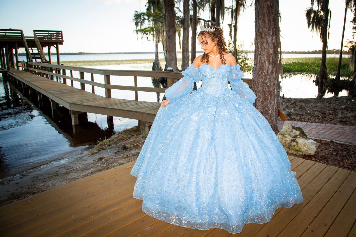 Young woman in a light blue ball gown quinceañera dress poses on a wooden dock near water surrounded by trees.