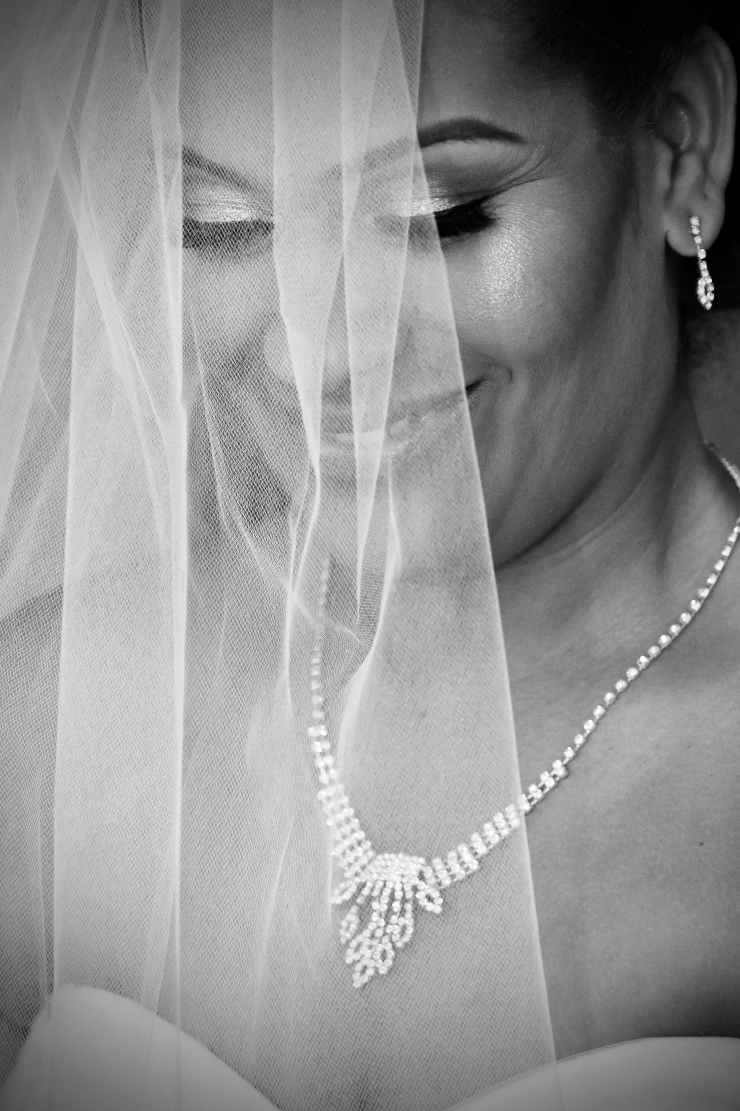 Black and white bridal portrait of a smiling bride with veil draped over face, wearing a sparkling diamond necklace and earrings.
