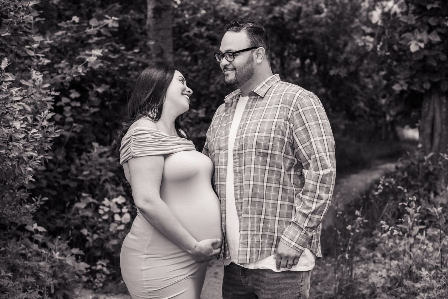 Black and white maternity photo of smiling couple outdoors, pregnant woman cradling baby bump, surrounded by lush foliage.