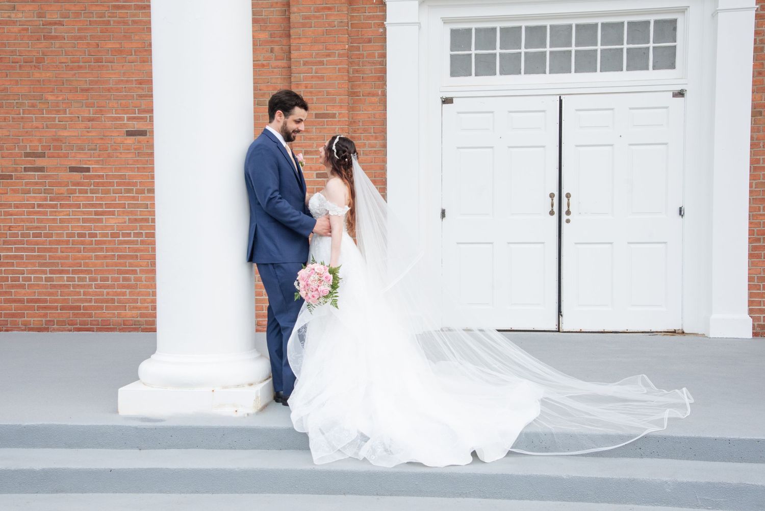 Bride and groom pose by white column outside brick church, her veil flowing gracefully in the breeze.
