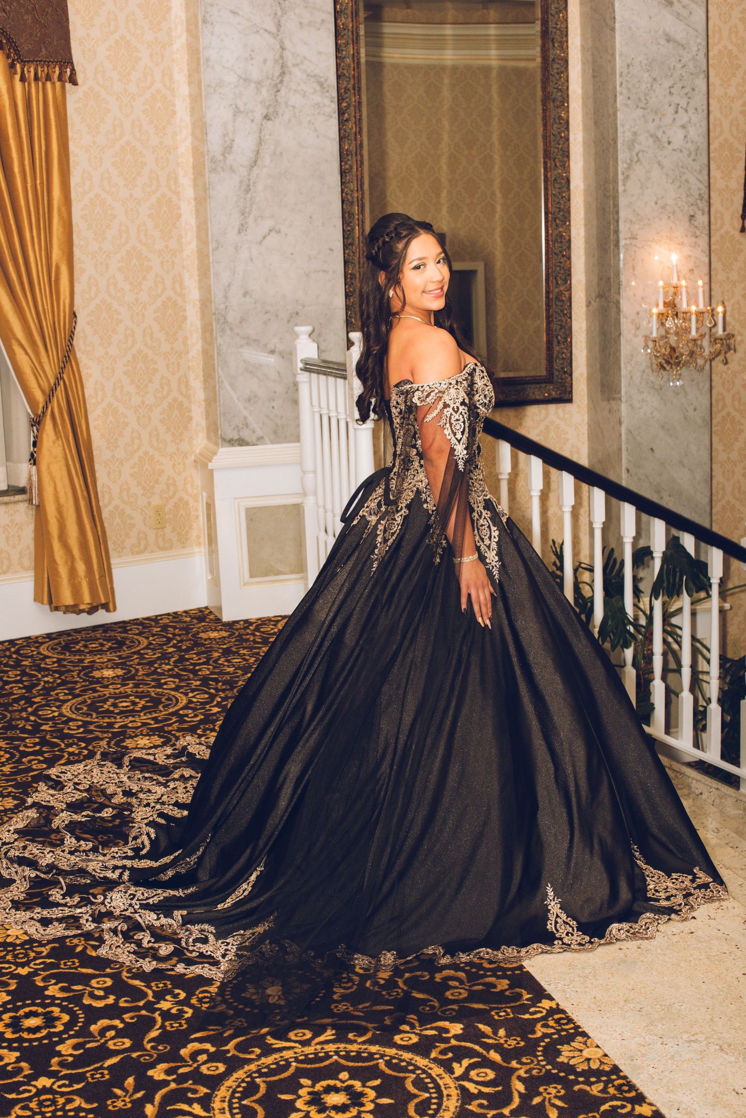Woman in elegant black and gold ball gown with sheer sleeves poses on grand staircase in ornate venue.
