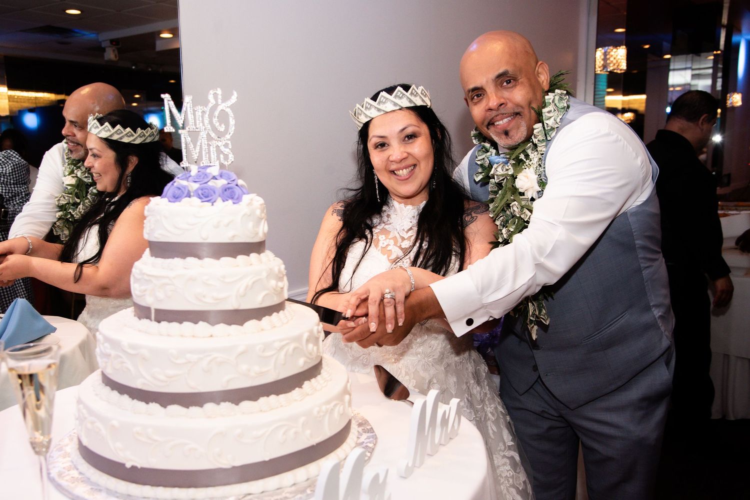Wedding guests gather around a white and silver tiered cake at an elegant reception celebration.