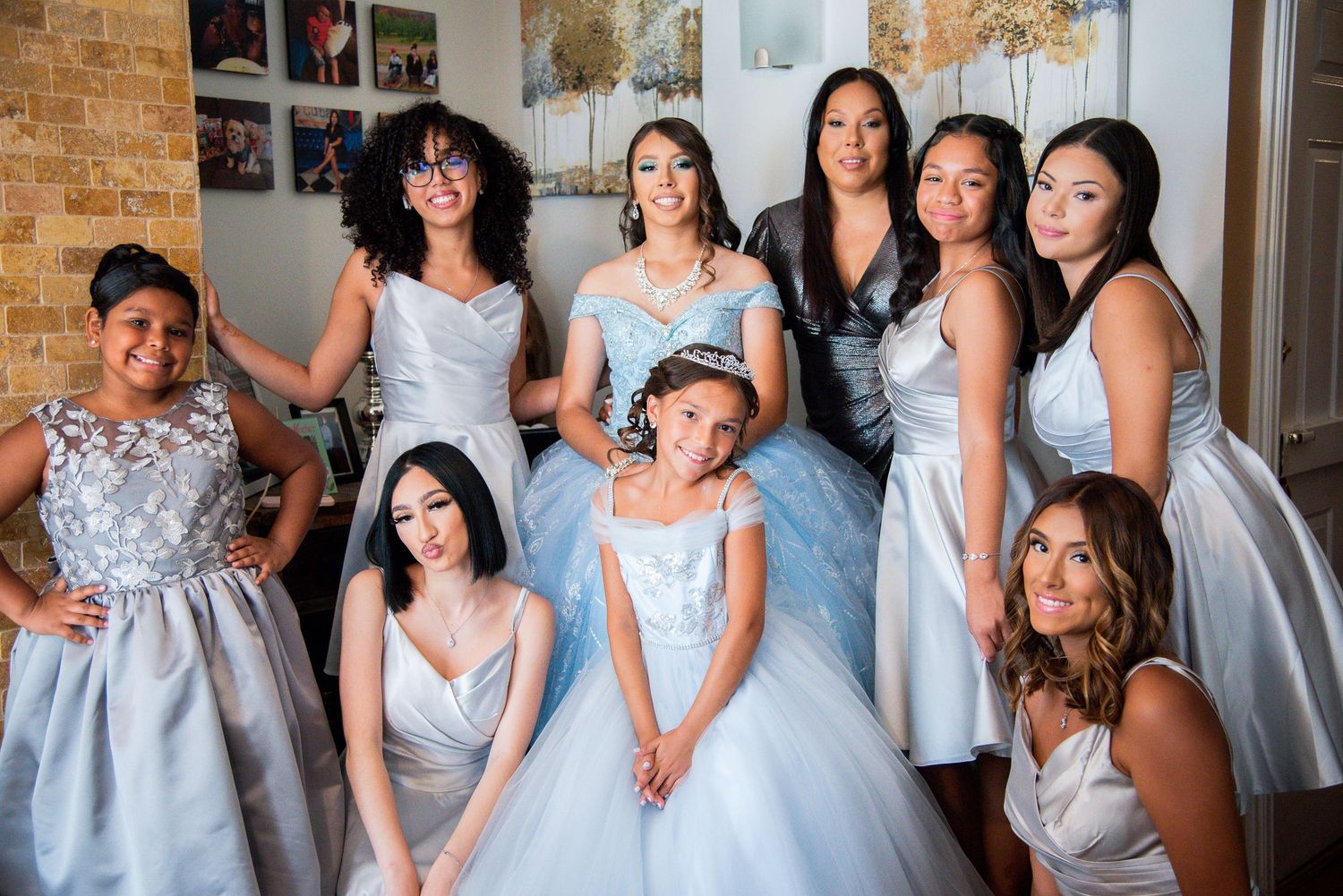 A quinceañera girl in a light blue ball gown poses with her court of honor in matching silver dresses indoors.