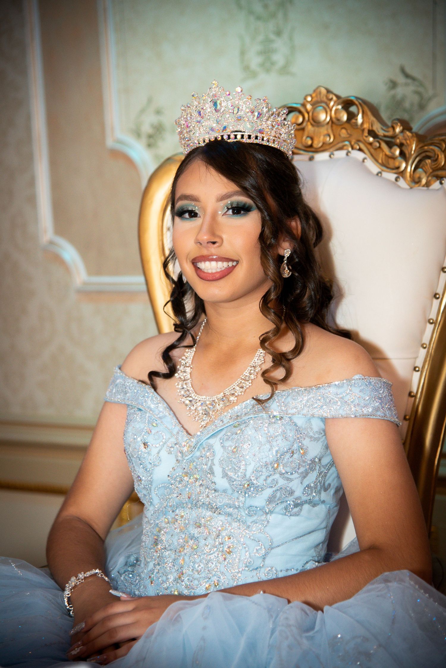 Young woman in light blue quinceañera gown and crystal crown sits on ornate gold throne chair, smiling elegantly by FBM Studios in Fort Laud