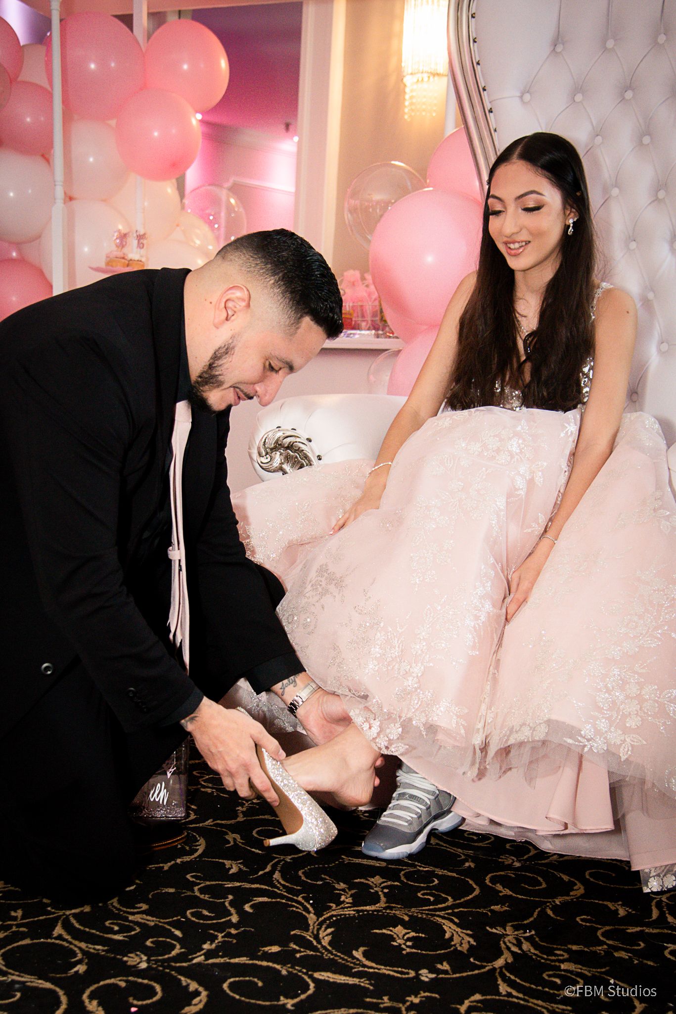 Man in black suit helps woman in pink ball gown with her shoe at a pink balloon-decorated celebration by FBM Studios in Fort Lauderdale Fl