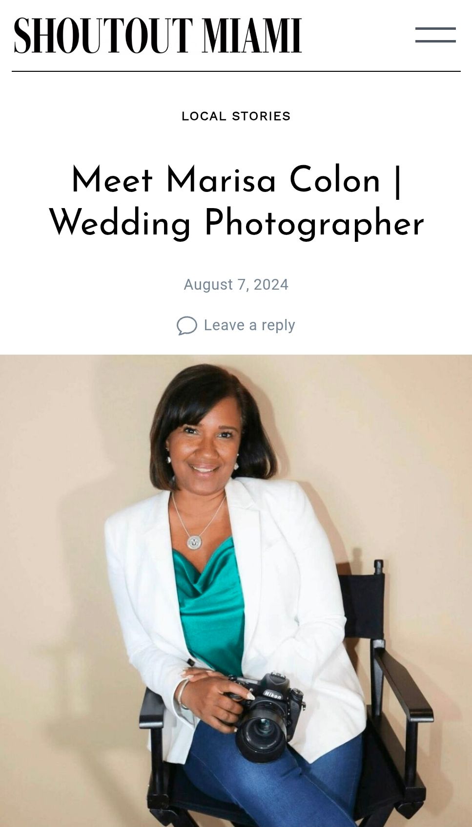 Professional photographer in white blazer and teal top holds camera while seated in studio setting by FBM Studios in Ft Lauderdale Fl.